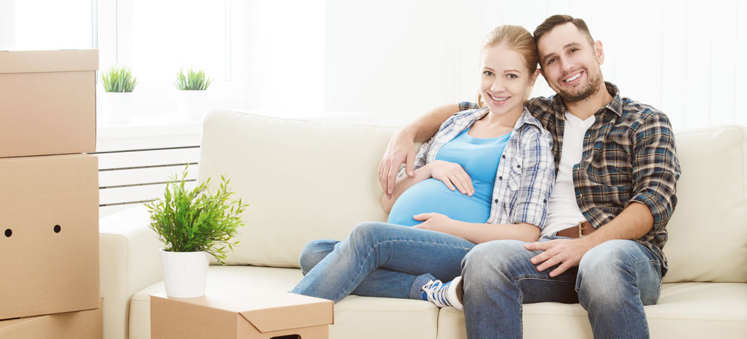 Pareja sentada en el sofá, la mujer embarazada, el hombre abrazándola con un brazo y cajas en el suelo